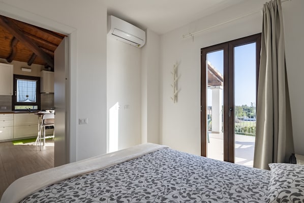 A bedroom with a bed, an air conditioning unit, a sliding glass door to a balcony, and a kitchenette in the adjacent room.