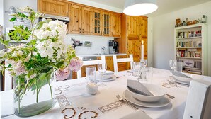 Un comedor con una mesa dispuesta para cuatro, armarios de madera, una estantería y un florero con flores.