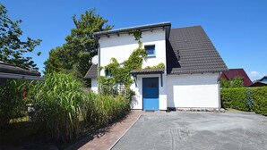 Una casa de dos pisos con una puerta azul, rodeada de follaje y un camino de entrada pavimentado.