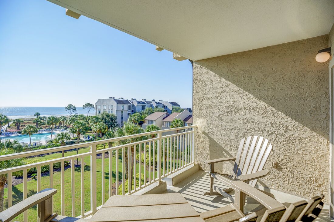 Un balcon offrant une vue sur une piscine et l’océan, entouré de verdure et d’un ensemble d’immeubles.