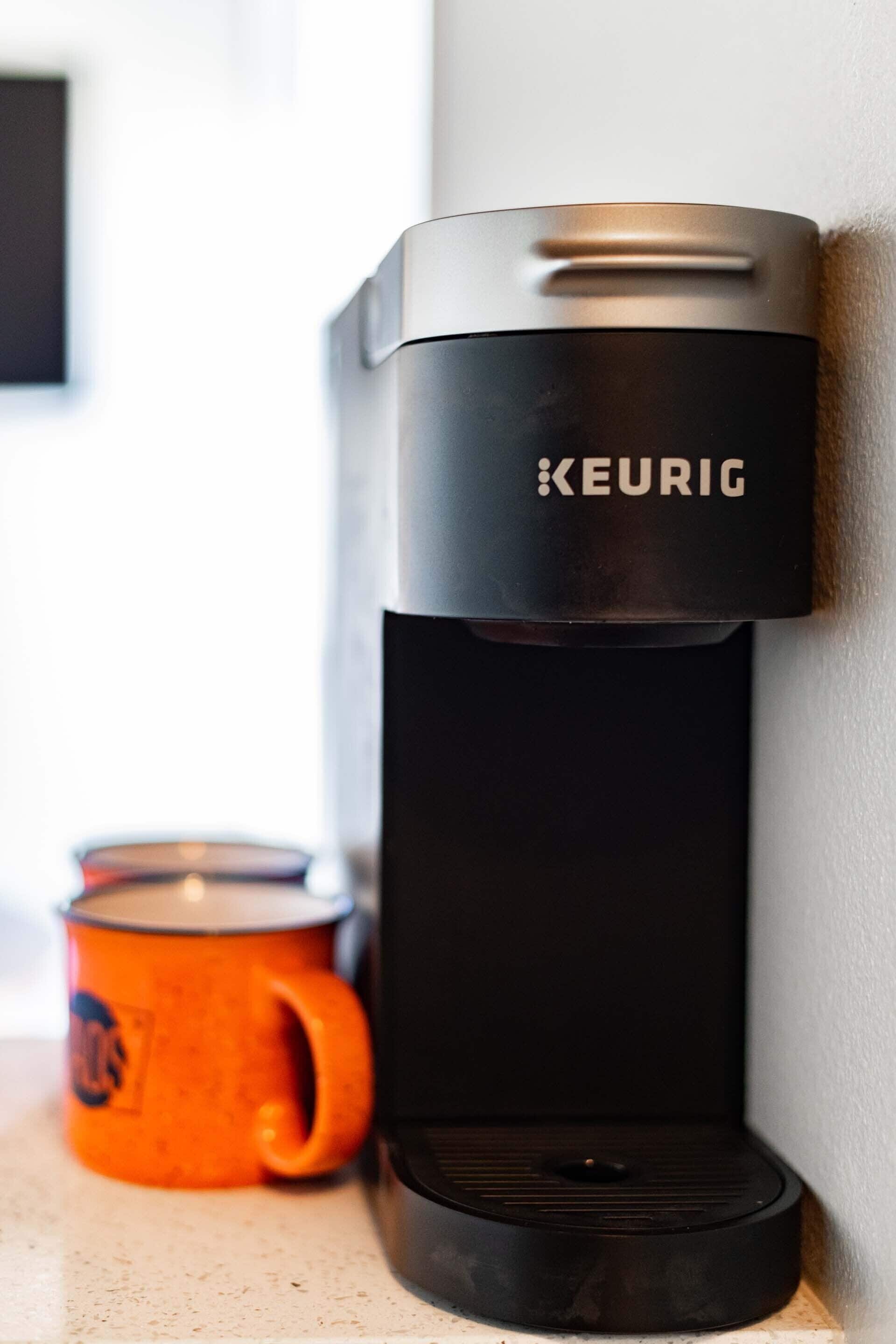 A black Keurig coffee machine mounted on a wall next to an orange mug.