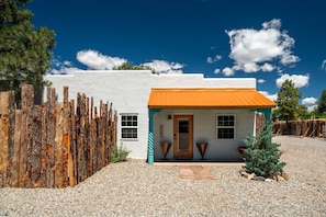 A white building with a wooden fence, a door, and a small porch with two chairs. - Luxx Motor Lodge (Ranchos De Taos)