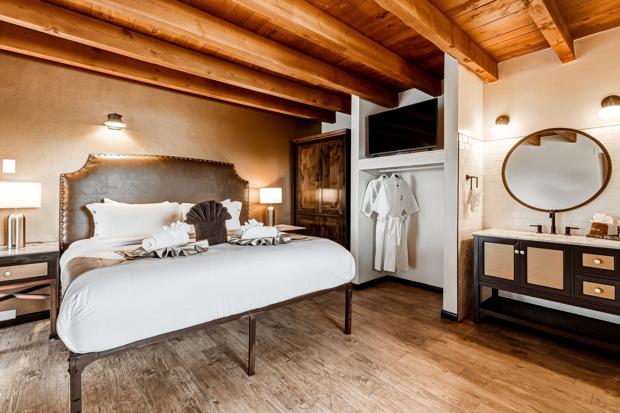 A hotel room with a large bed, wooden ceiling beams, a TV, a mirror, and a bathroom with a sink and towel rack.