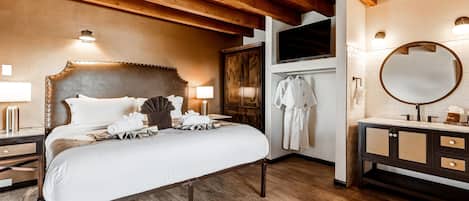 A hotel room with a large bed, wooden ceiling beams, a TV, a mirror, and a bathroom with a sink and towel rack.
