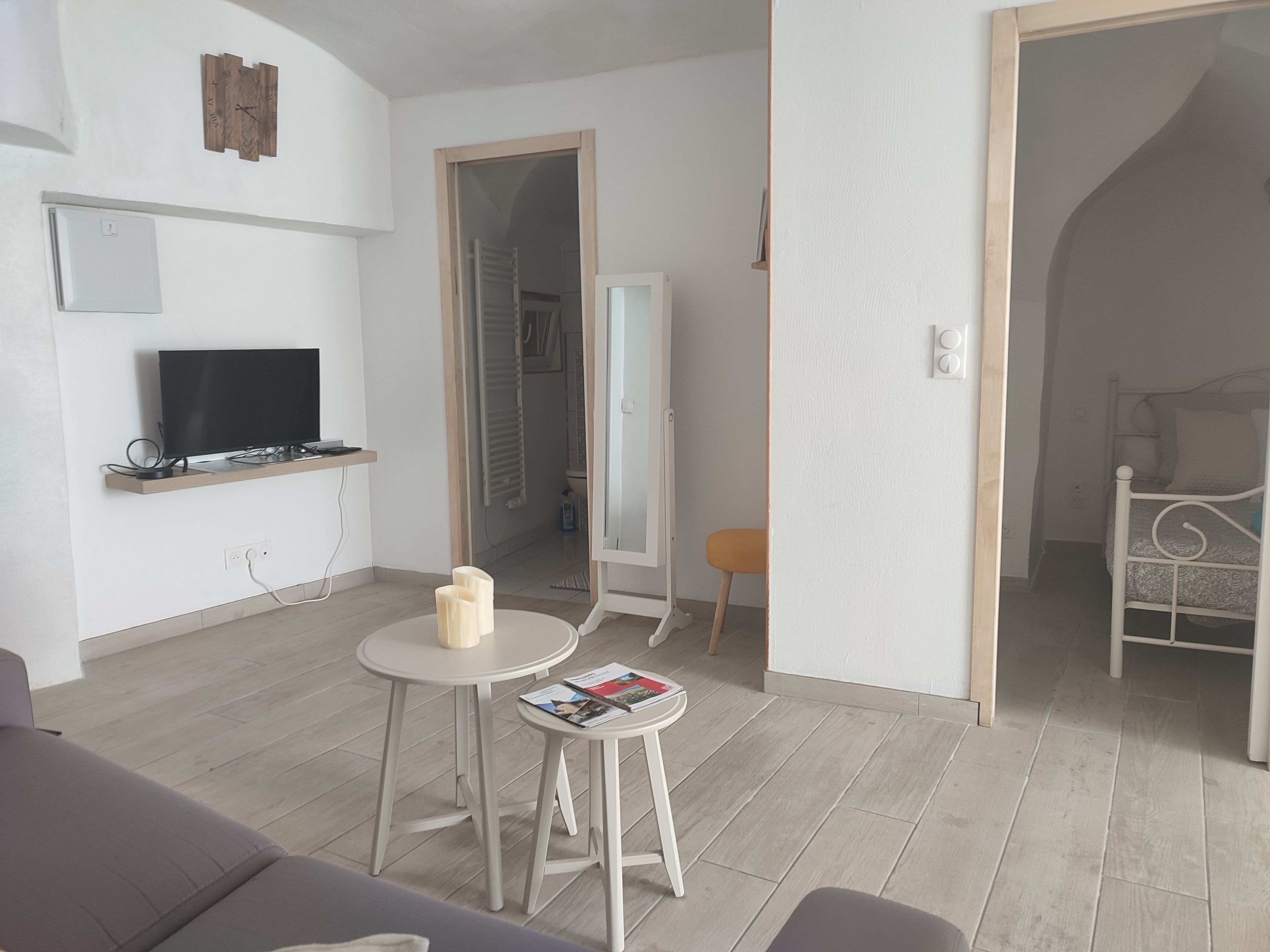 A modern living room with a flat-screen TV, a small round table with magazines, a sofa, and a bed in the adjacent room.