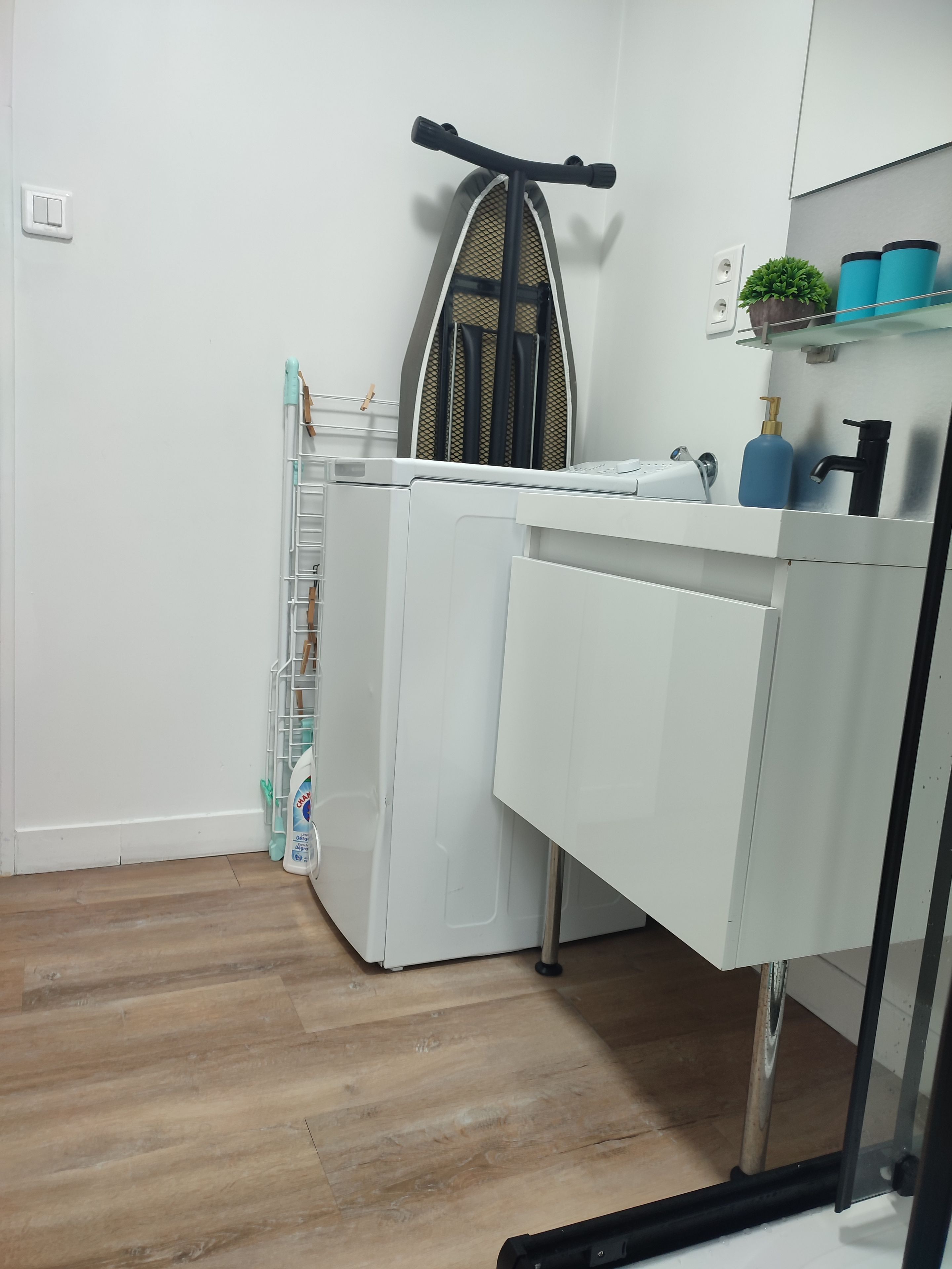 A compact laundry room with a washing machine, drying rack, and a small shelf with toiletries.