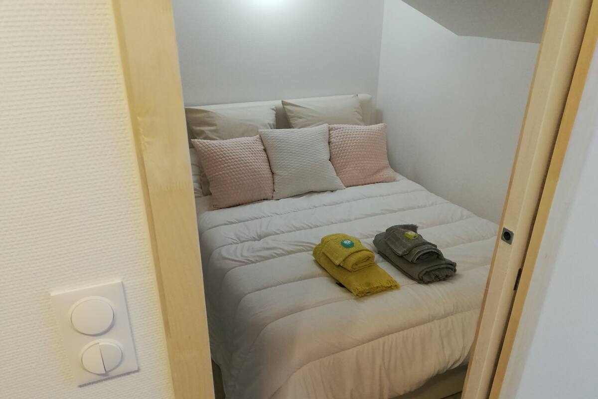 A neatly made bed with a white duvet, multiple pillows, and two rolled towels on the bed.