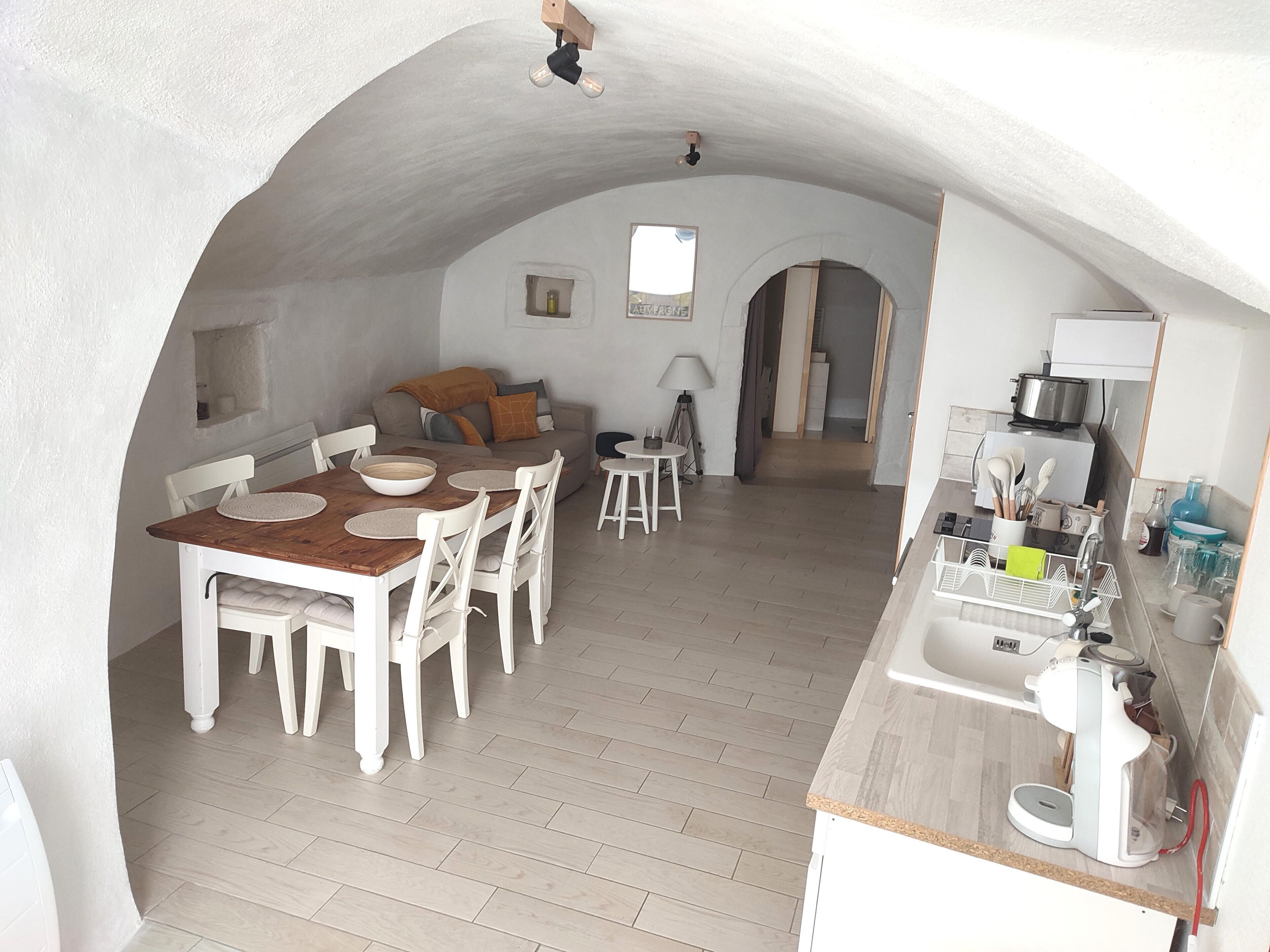 A cozy, open-plan living and dining area with a wooden dining table, white chairs, and a sofa. A kitchen area with modern appliances is visible in the background.