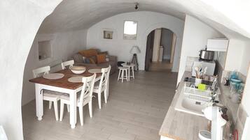 A cozy, open-plan living and dining area with a wooden dining table, white chairs, and a sofa. A kitchen area with modern appliances is visible in the background.