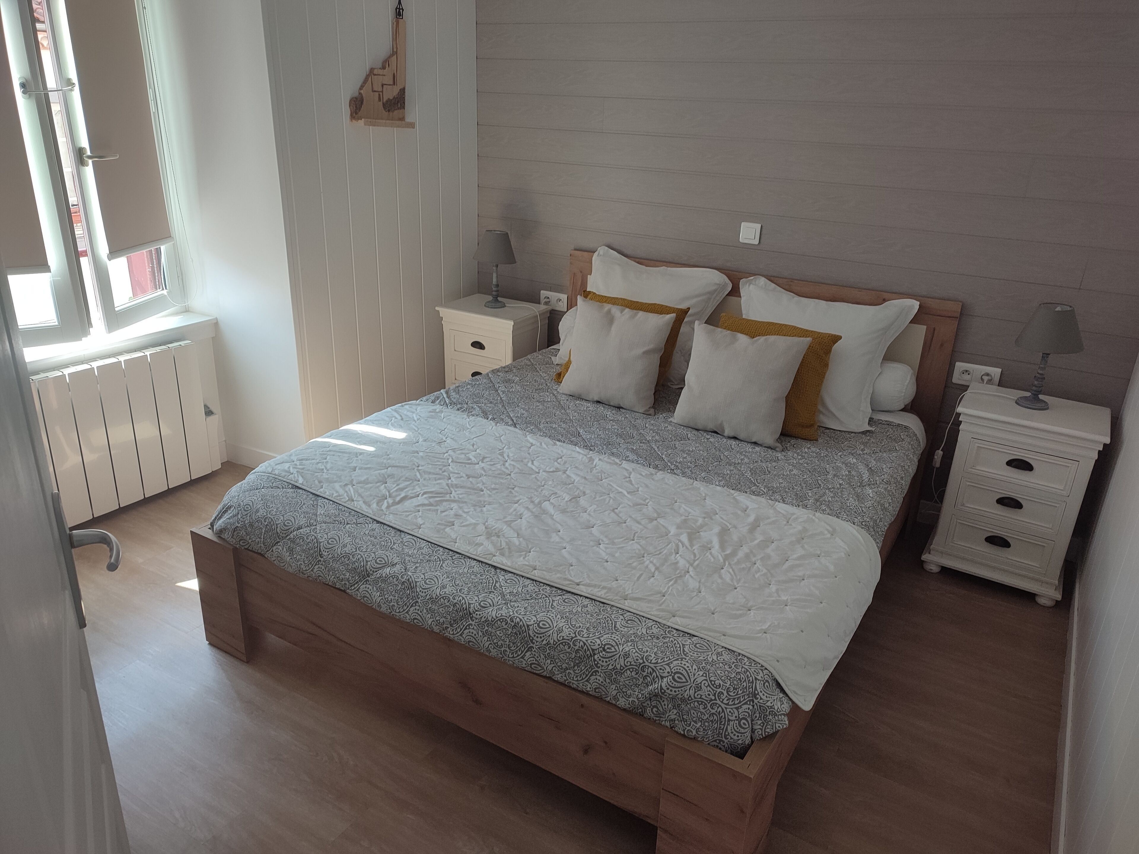 Une chambre à coucher avec un lit en bois, une literie blanche, deux tables de chevet avec des lampes, un radiateur et une fenêtre avec des stores.