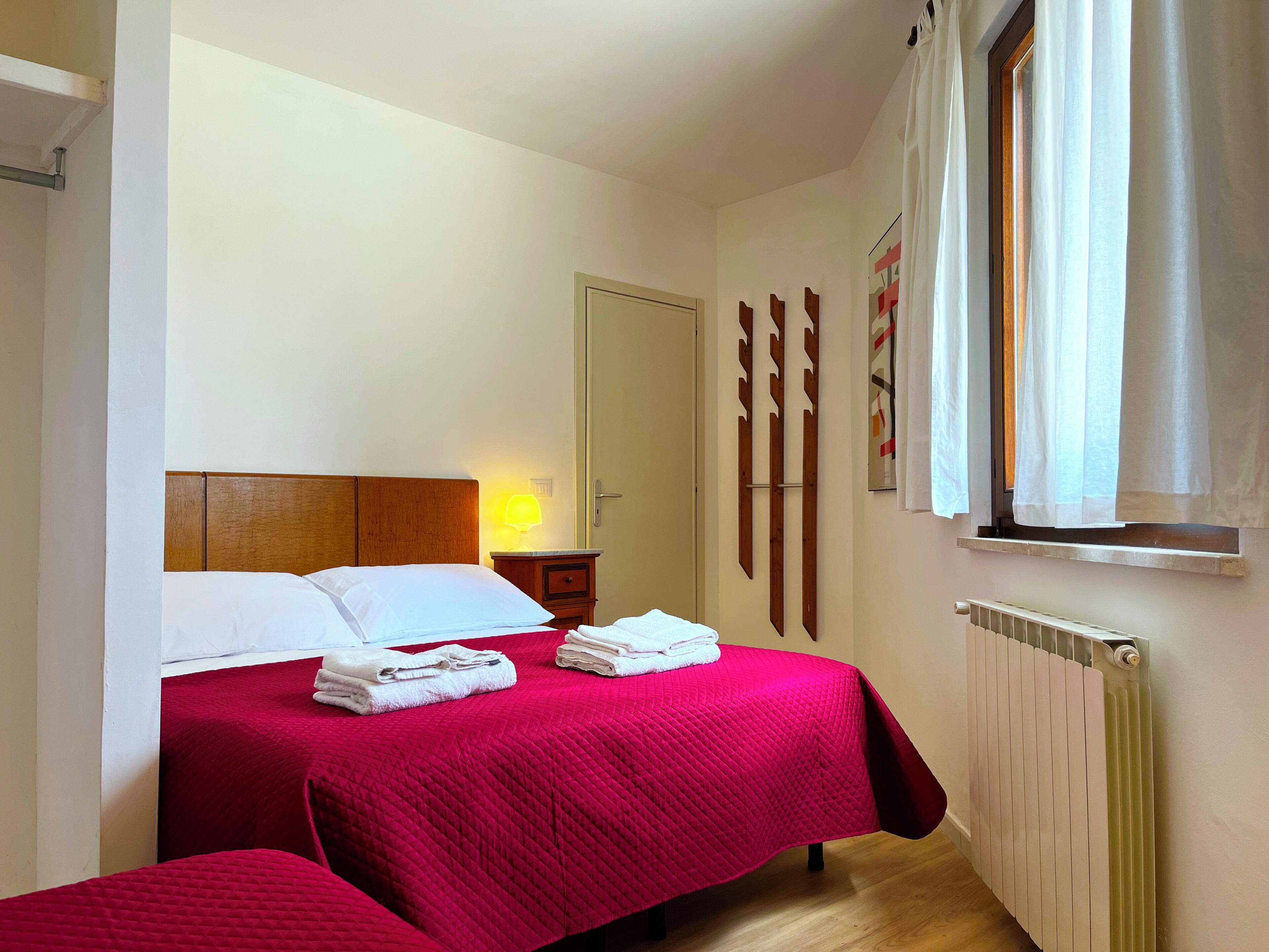A bedroom with two beds, a nightstand, a lamp, a radiator, and a window with curtains.