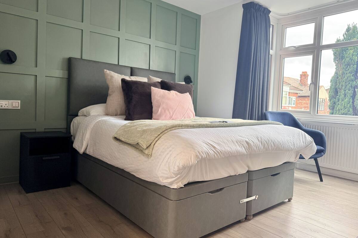 A modern bedroom with a grey bed, a nightstand, a chair, and a window with a view of buildings.