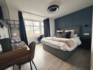 A modern hotel room with a large bed, a desk with water bottles, a chair, and a view of the outside.