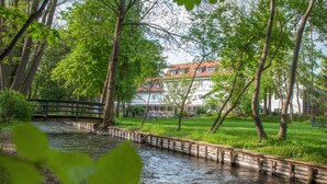 Une rivière traversée par un pont en bois, entourée d’une végétation luxuriante, avec un bâtiment au toit rouge en arrière-plan.