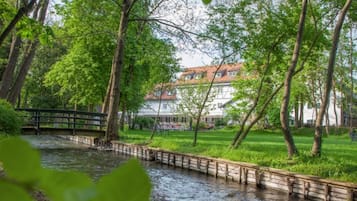 A riverside walkway with a wooden bridge, surrounded by lush greenery and a building with a red roof in the background.