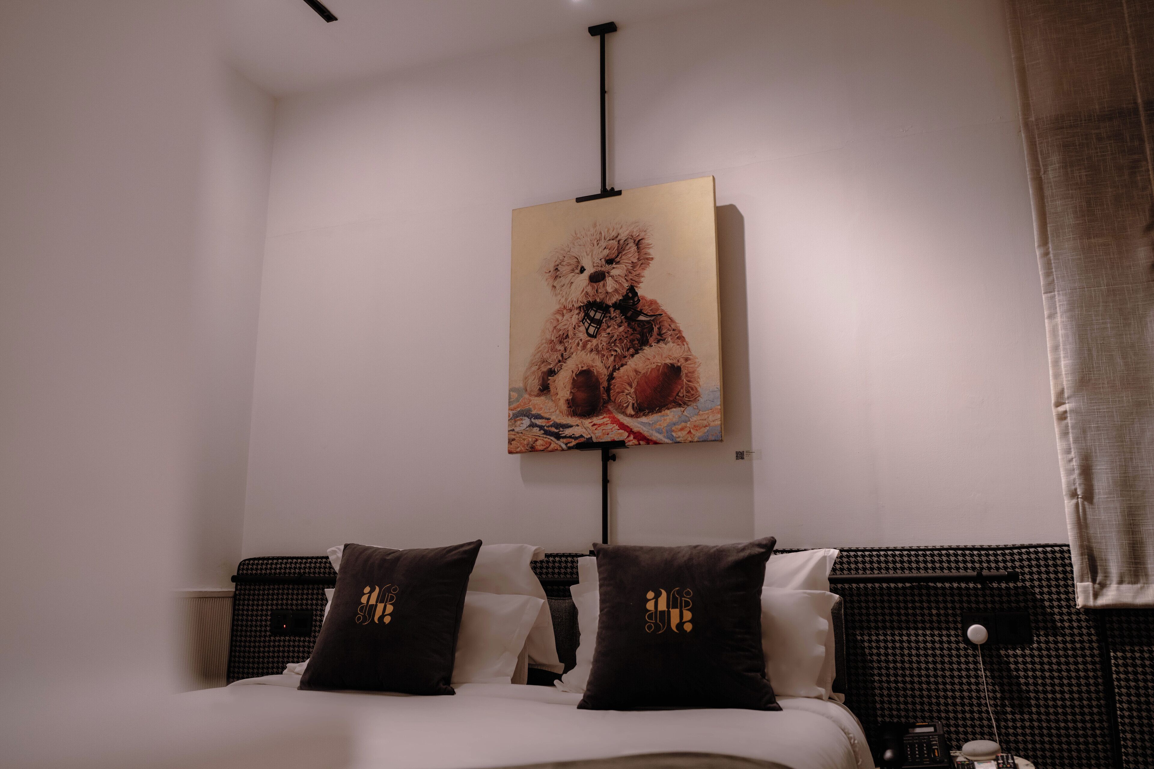 Quarto com cama, duas almofadas com capas bordadas, um quadro na parede de um urso de pelúcia e uma mesa de cabeceira com abajur.