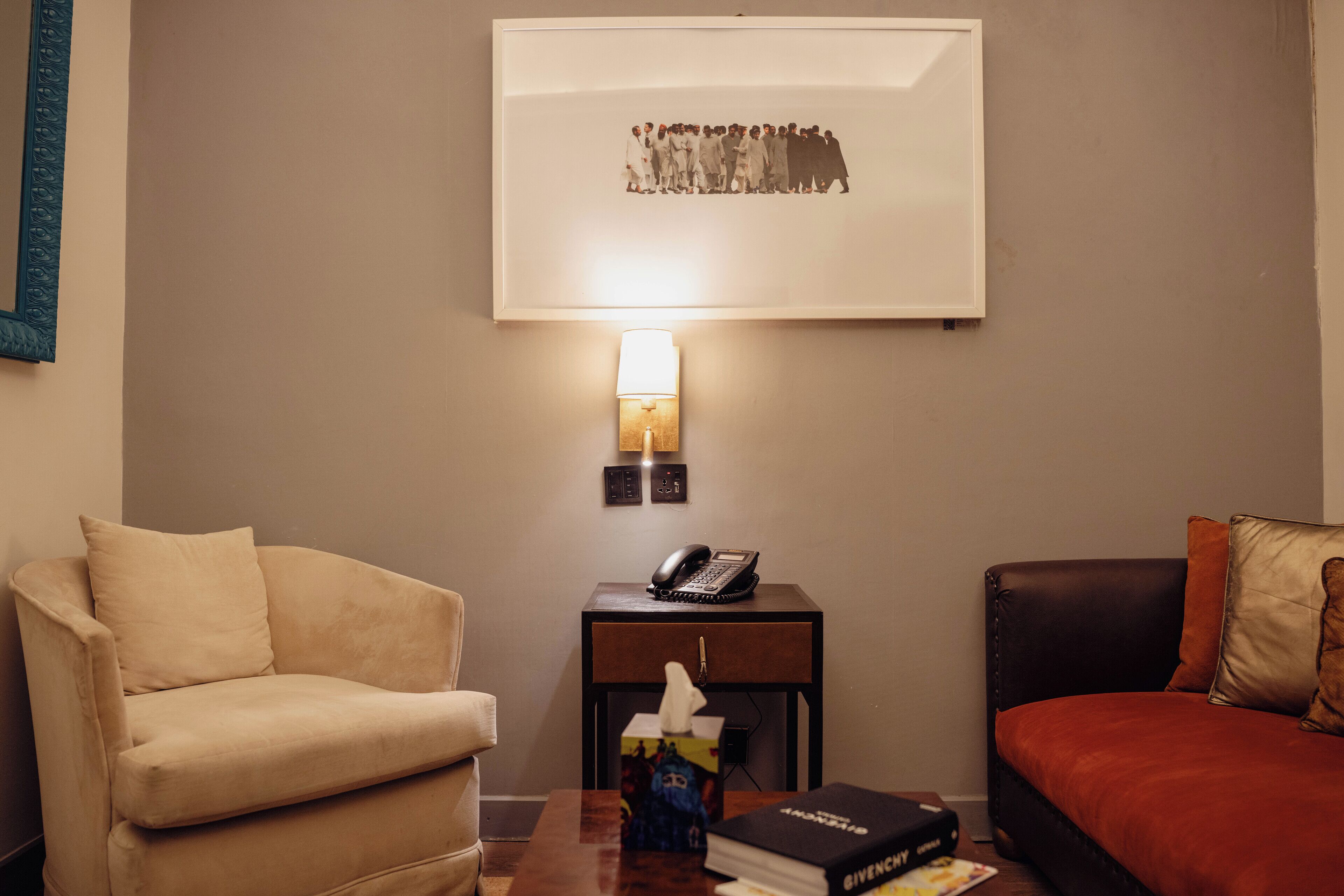 A hotel room with a beige armchair, a small bedside table with a lamp, a telephone, a tissue box, and a book.
