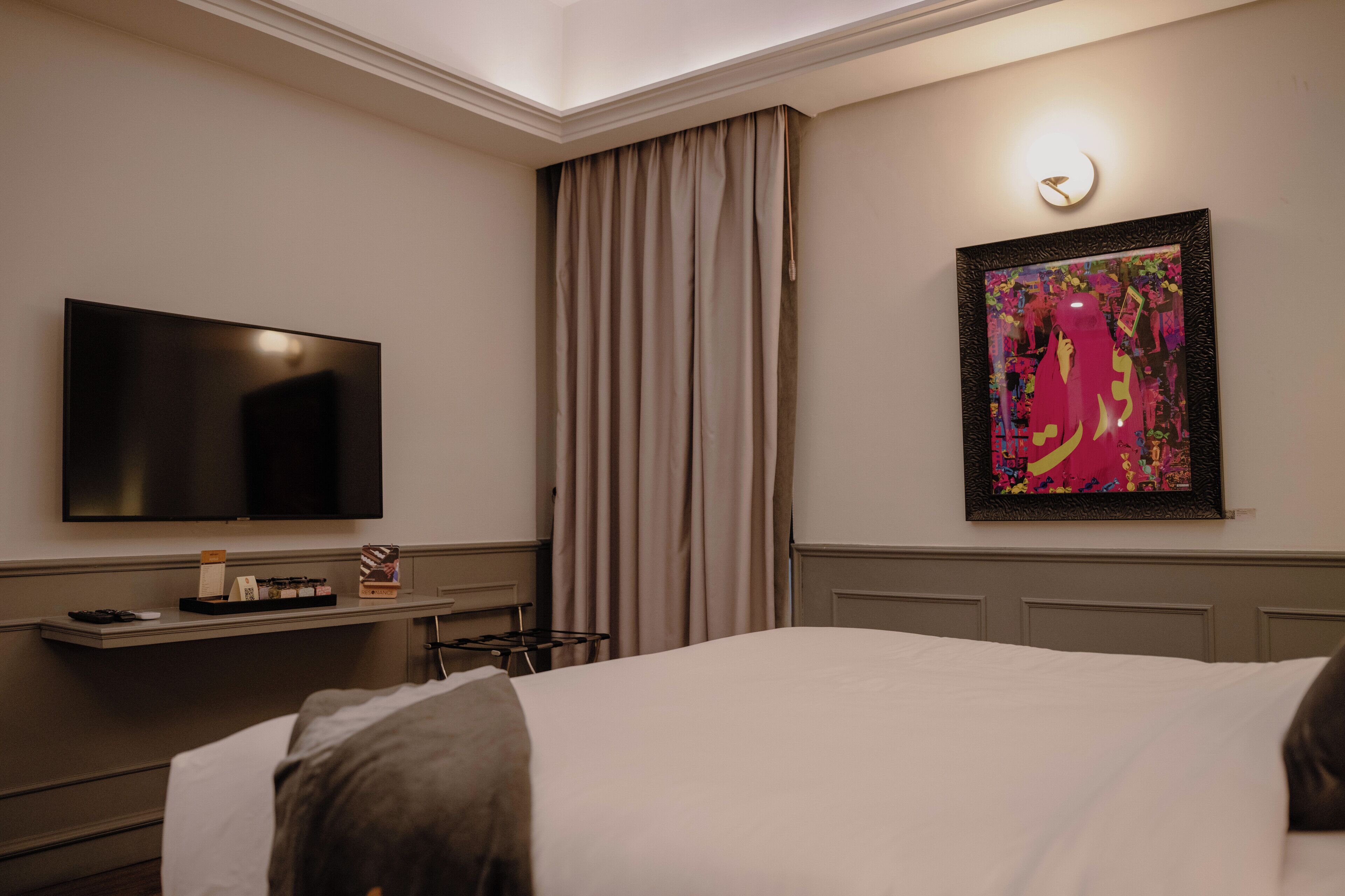 Habitación de hotel con cama, televisor de pantalla plana, escritorio con lámpara, un cuadro en la pared y ventana con cortinas.