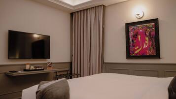 Habitación de hotel con cama, televisor de pantalla plana, escritorio con lámpara, un cuadro en la pared y ventana con cortinas.