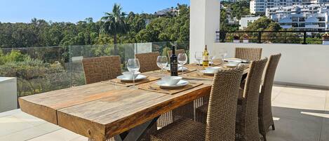 Een houten eettafel gedekt met witte borden, wijnglazen en een fles wijn op een balkon met rieten stoelen.
