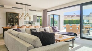 Een moderne woonkamer met een bank, een salontafel en een eethoek met een tafel en stoelen. Een groot raam biedt uitzicht op de omgeving.