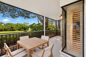 A balcony with a wooden table and chairs, a glass door with blinds, and a view of a garden with trees and plants.