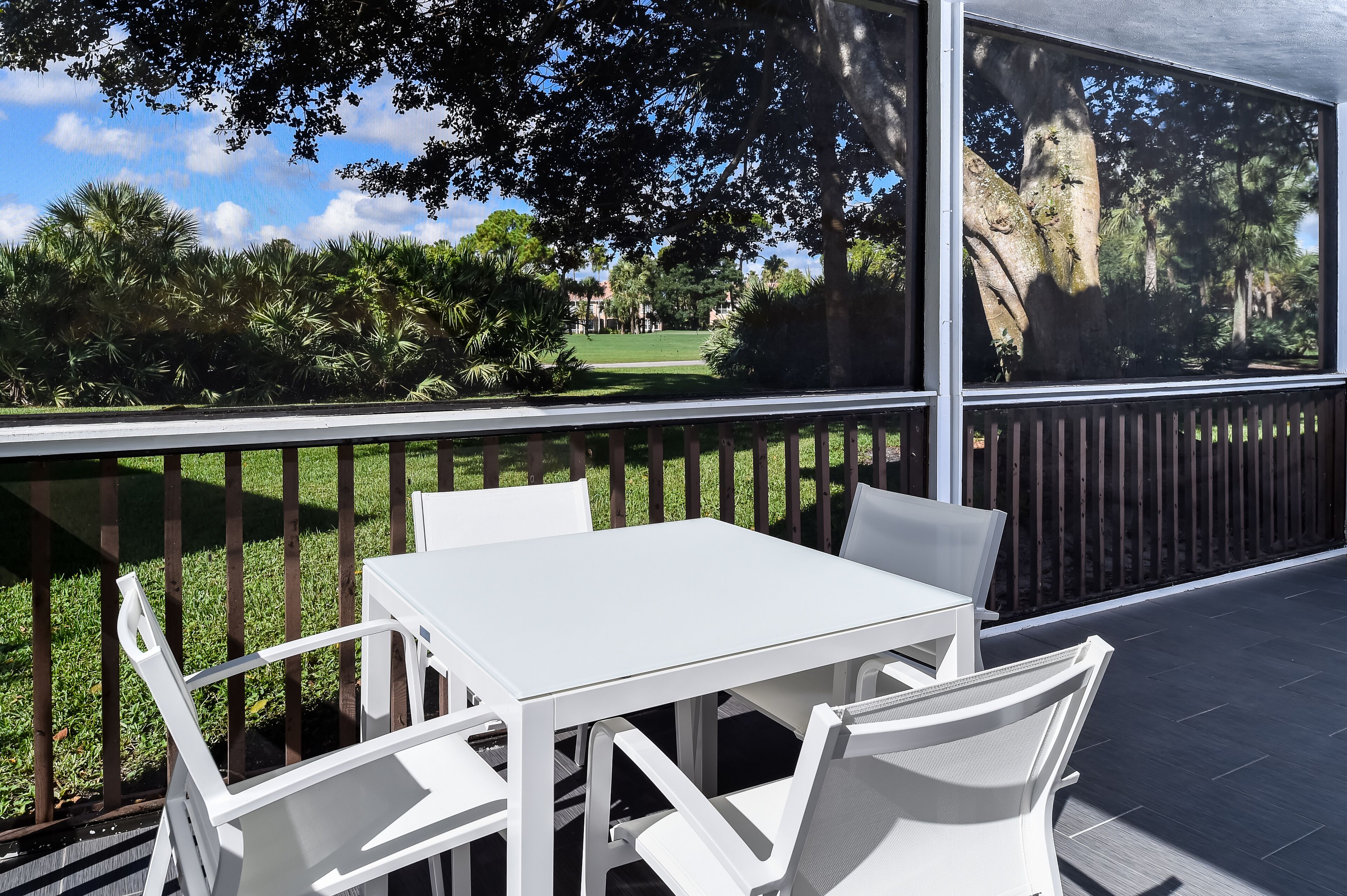 A balcony with a white table and chairs, overlooking a grassy area with trees and a distant building.