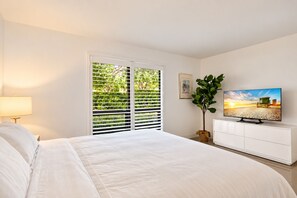 Ein Schlafzimmer mit einem Bett, einem Fernseher, einer Pflanze und einem Fenster mit Jalousien.