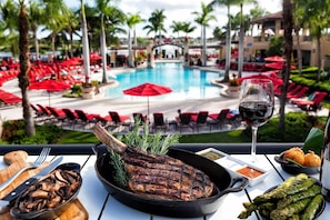 Ein Esstisch mit einem Steak, Spargel und einem Glas Rotwein, mit Blick auf einen Poolbereich mit roten Sonnenschirmen und Liegestühlen.