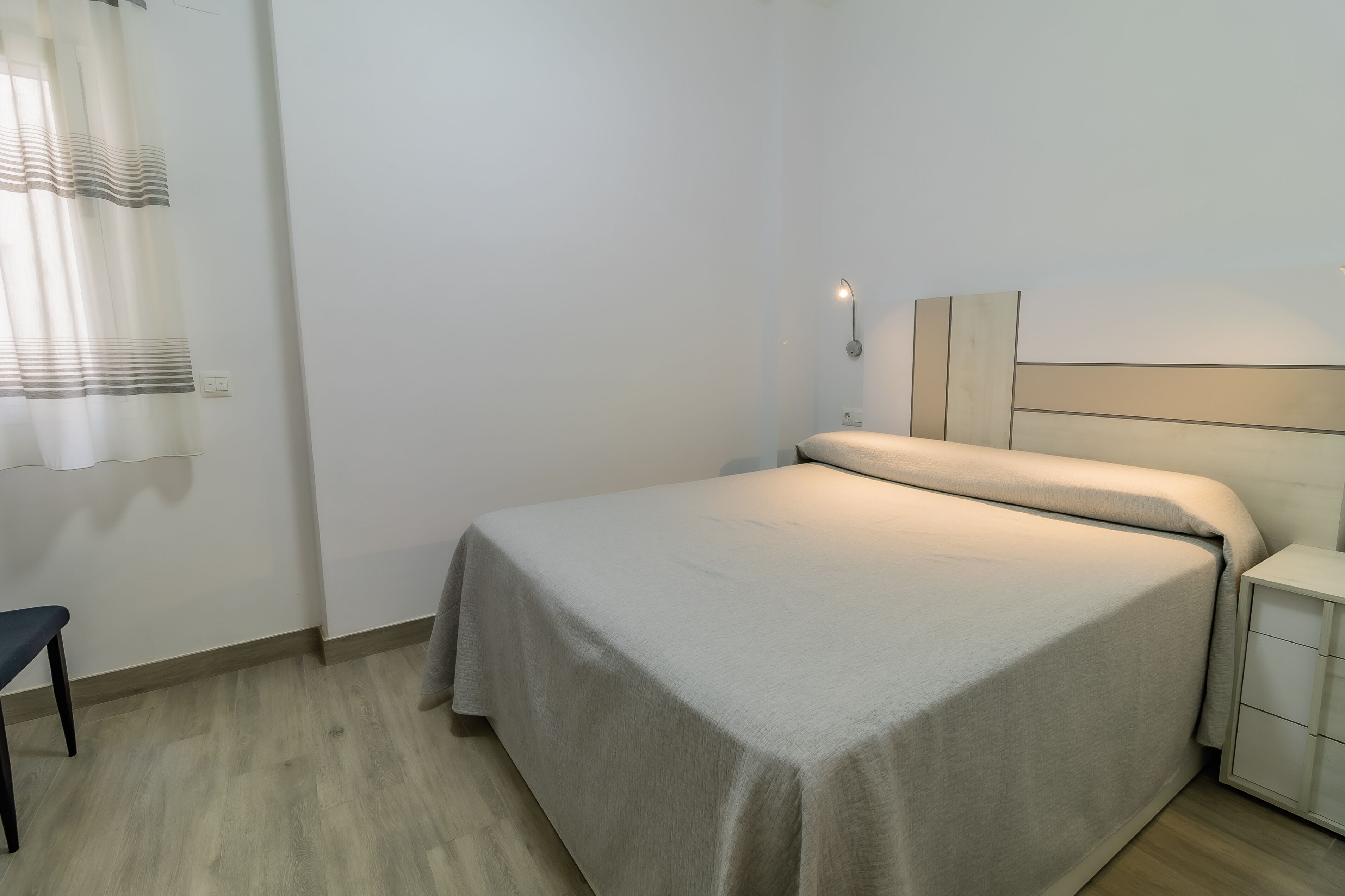 A neatly made bed with a light-colored bedspread, a bedside table with a lamp, and a chair.