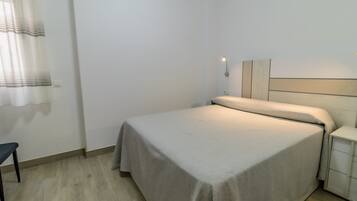 A neatly made bed with a light-colored bedspread, a bedside table with a lamp, and a chair.