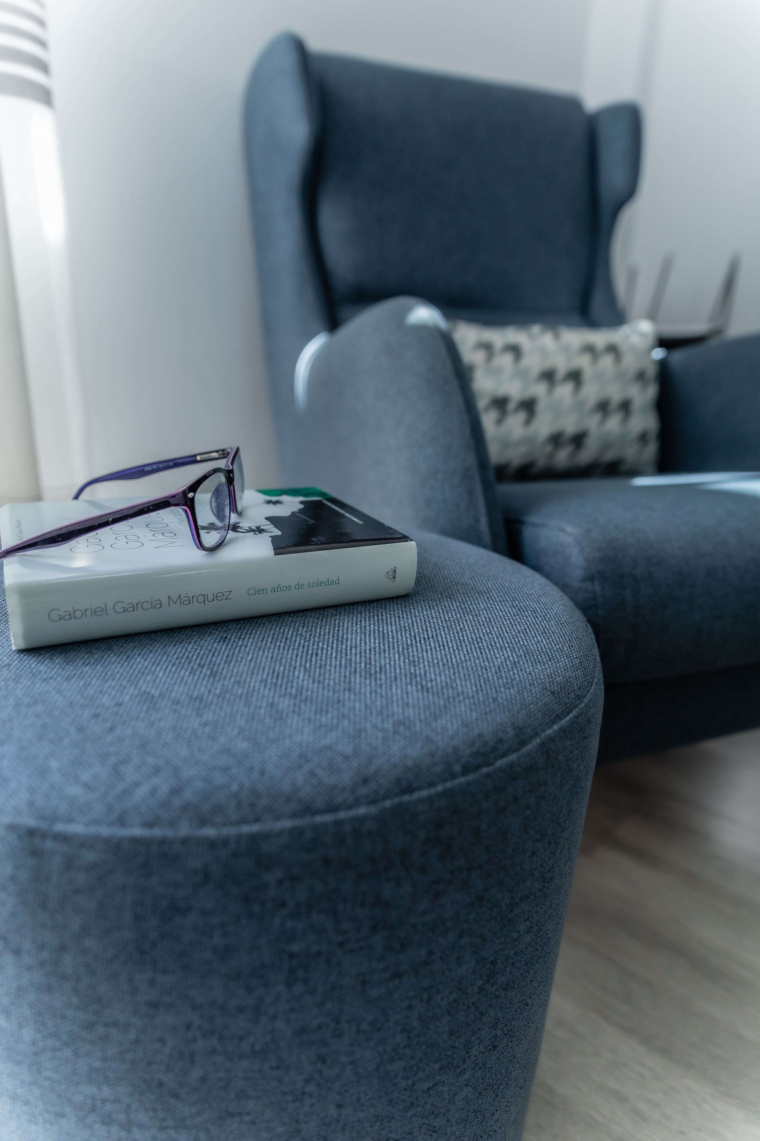 A book titled 'Cien años de soledad' by Gabriel Garcia Marquez, a pair of glasses, and a grey armchair with a patterned pillow in a modern living room.