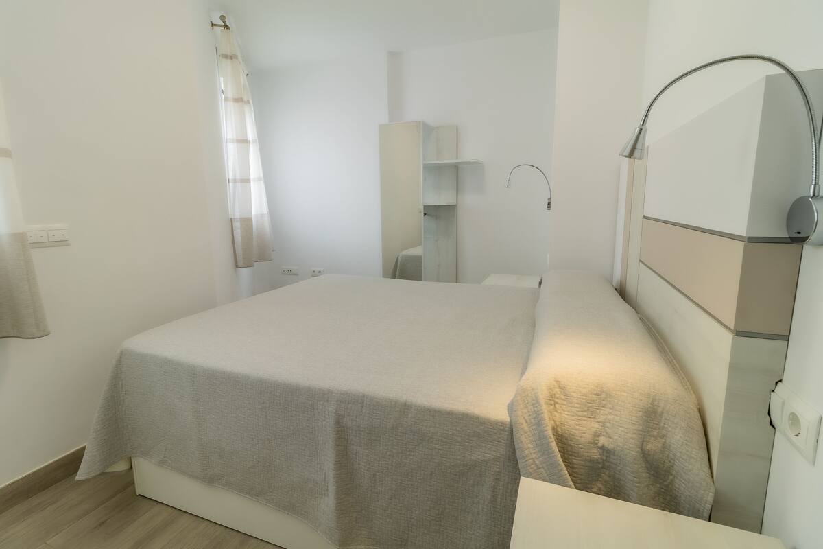 Een keurig opgemaakt bed met een grijze spreideken, een nachtkastje met een lamp en een wandplank.