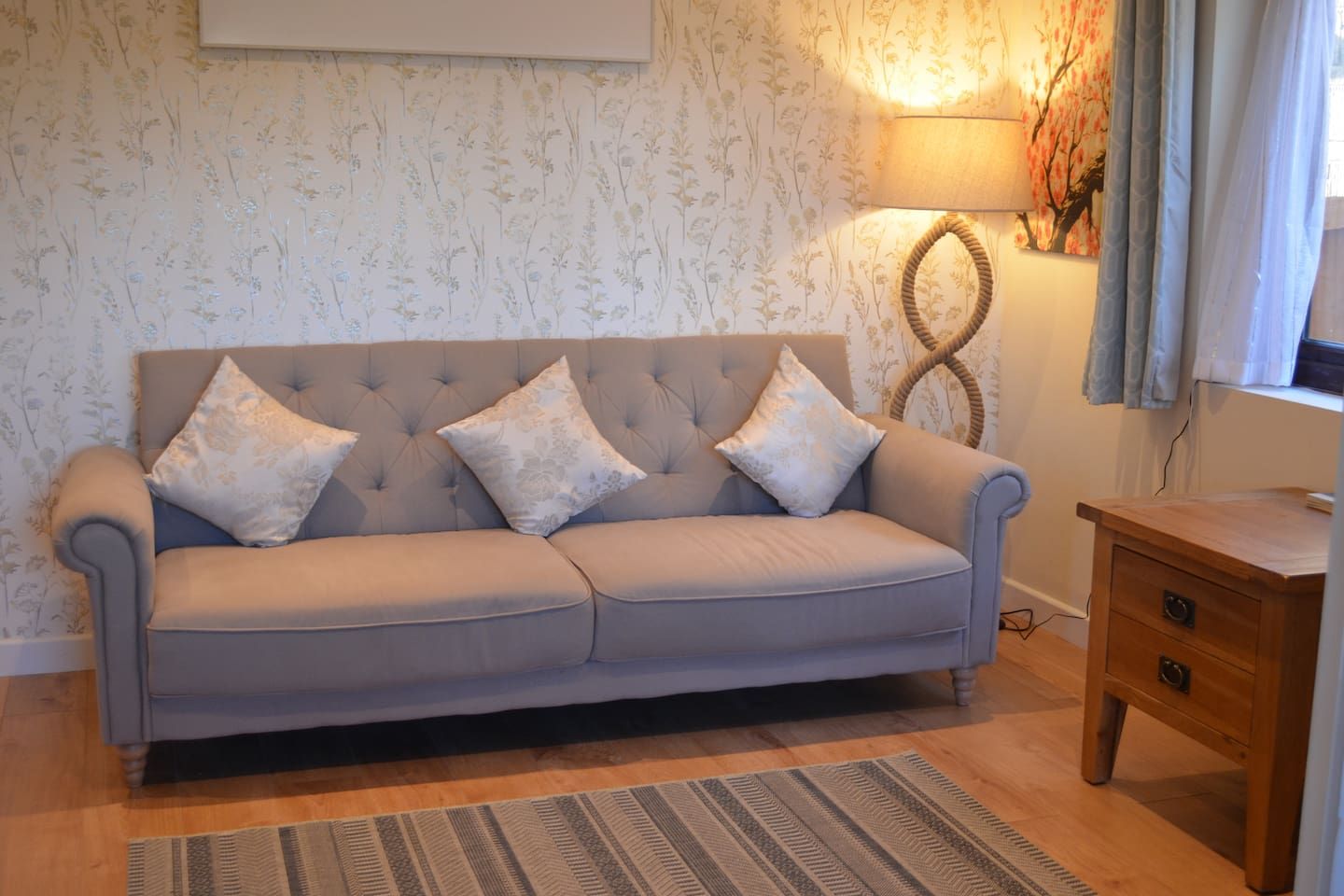 A sofa with floral cushions, a lamp, and a wooden side table in a room with floral wallpaper.