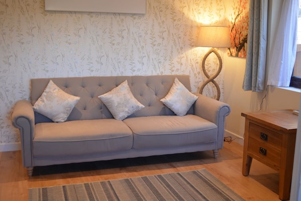 Een bank met bloemenkussens, een lamp en een houten bijzettafeltje in een kamer met bloemenbehang.