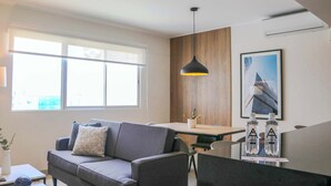 A modern living room with a gray sofa, a dining table, and a pendant light. There are two water bottles on the table.