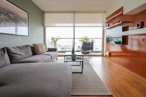 A modern living room with a grey sofa, a black chair, a coffee table, and a TV mounted on a wooden wall. - w* | 3BR Premium Cityview w/ Balcony in Miraflores (Miraflores)