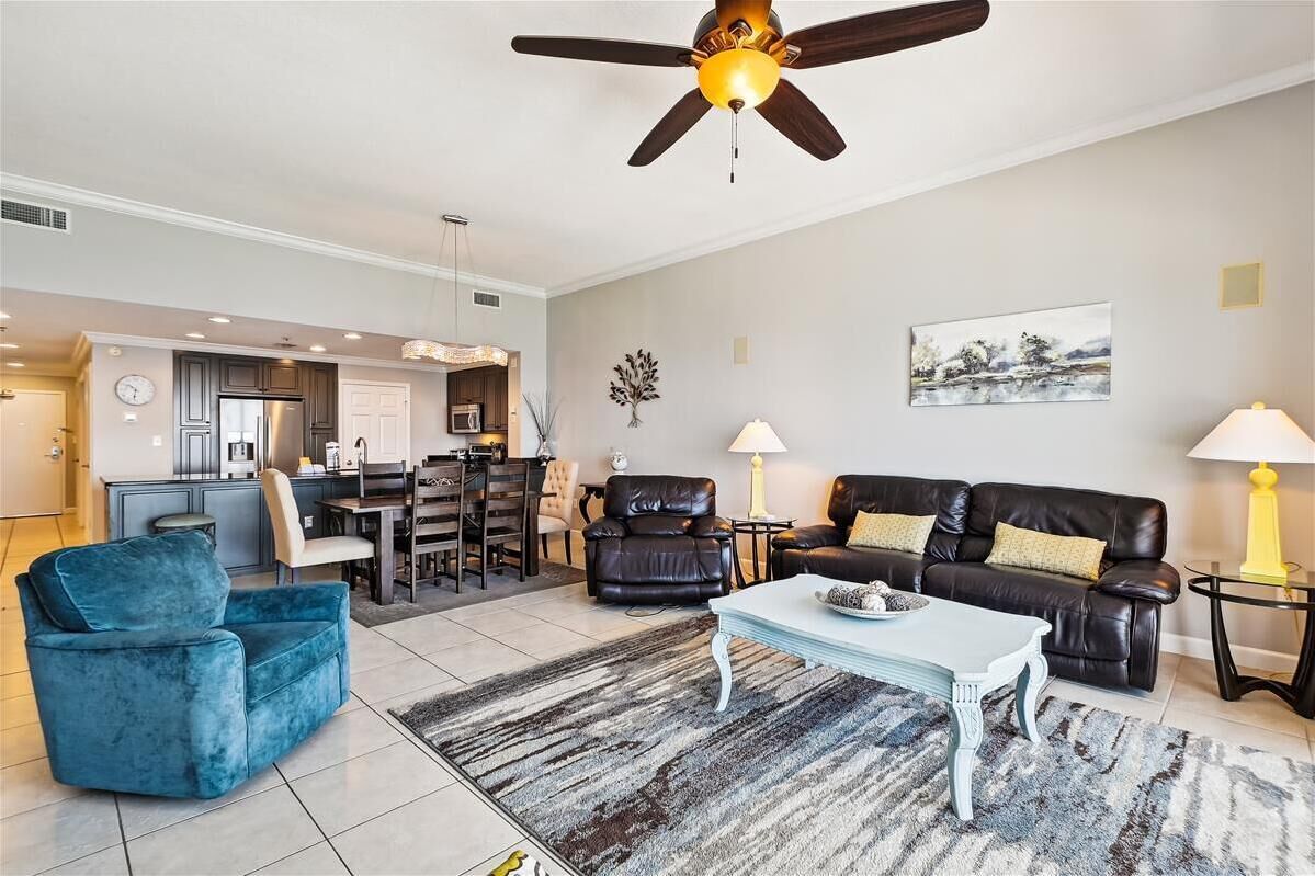 A modern living room with a black leather sofa, a blue armchair, and a white coffee table. There is a ceiling fan, a dining area with a table and chairs, and a kitchen in the background.