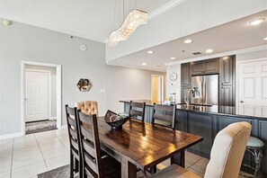 A dining area with a wooden table and chairs, a chandelier, and a kitchen with cabinets and a refrigerator.