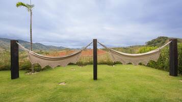 A hammock set up between two wooden posts in a grassy area with a view of hills and a palm tree in the background.