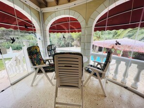 A covered patio area with a table and chairs, large windows with red awnings, and a view of greenery outside.