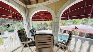 A covered patio area with a table and chairs, large windows with red awnings, and a view of greenery outside. - T-0622, Evelyn, villa with private pool in Dènia (Denia)