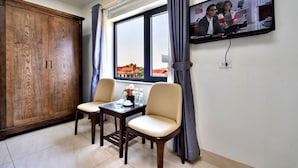 Una habitación con un armario de madera, dos sillas, una mesita con un florero y botellas de agua, una ventana con cortinas y un televisor montado en la pared.