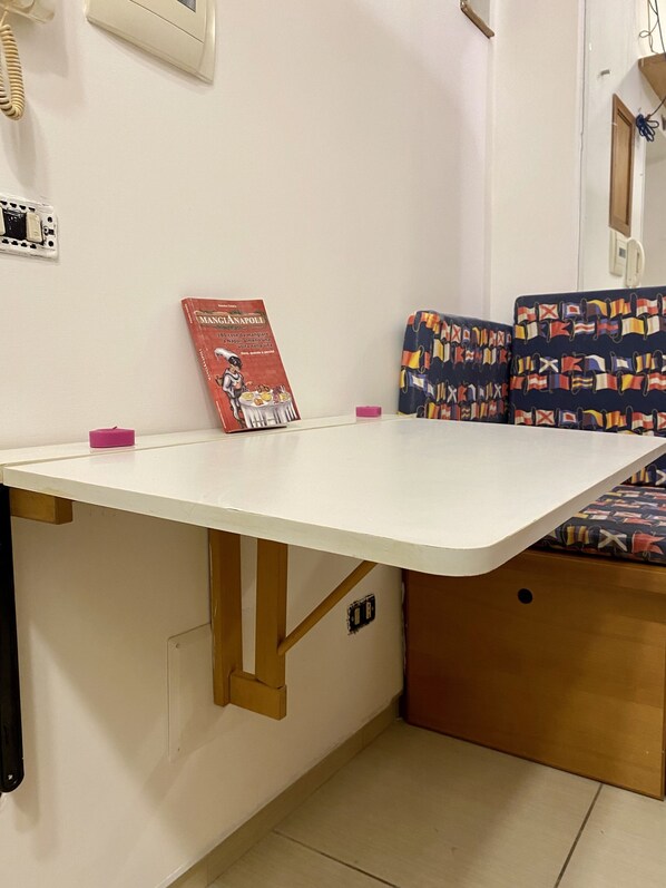 A small, compact room with a white table, a red book titled 'MangiaNapoli', and a bench with a flag pattern. - Boat Apartment (Napoli)