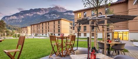 An outdoor patio with tables and chairs, a wooden pergola, and a mountain backdrop.