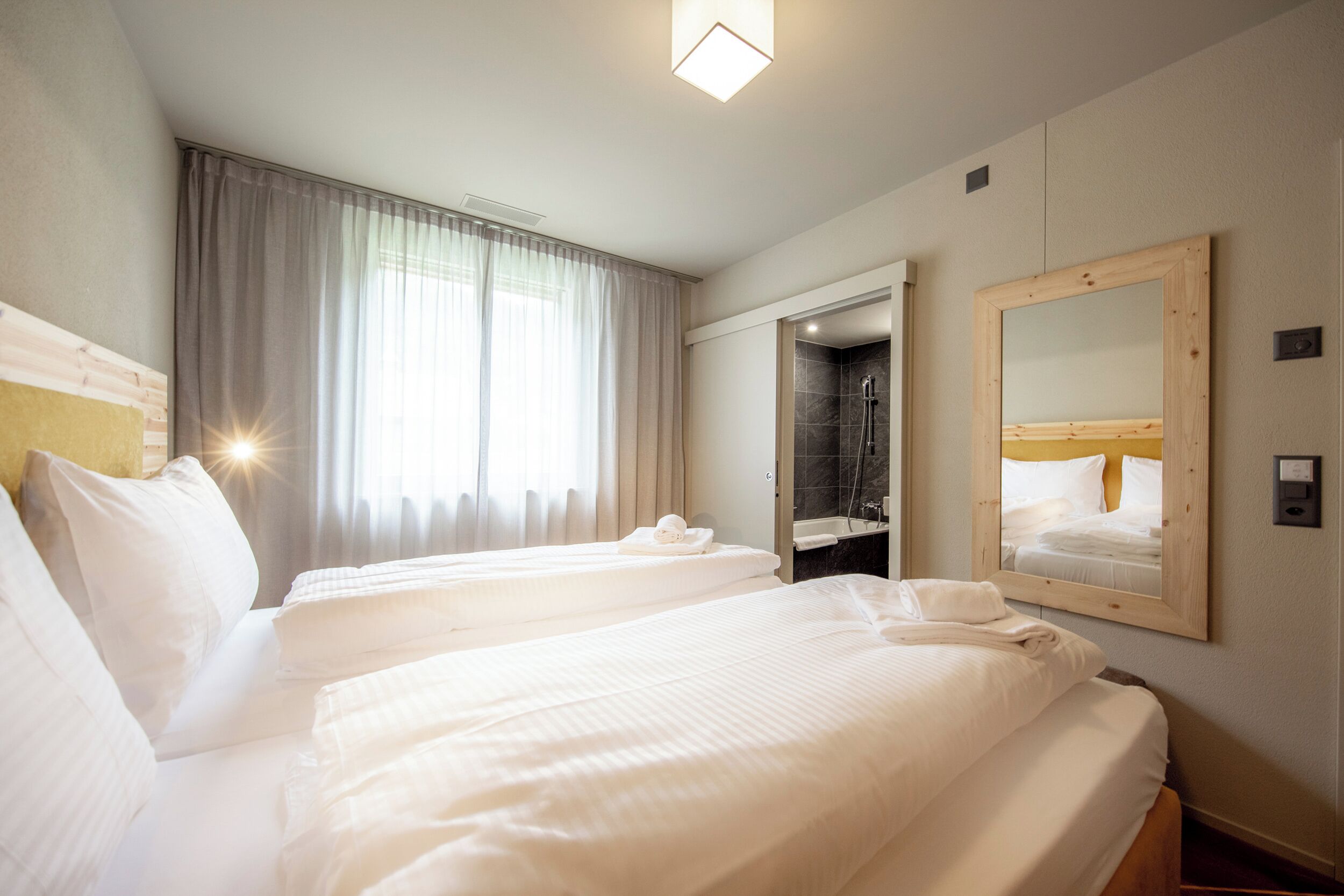 Una camera d'albergo con due letti, bagno con doccia, uno specchio e una finestra con tende.