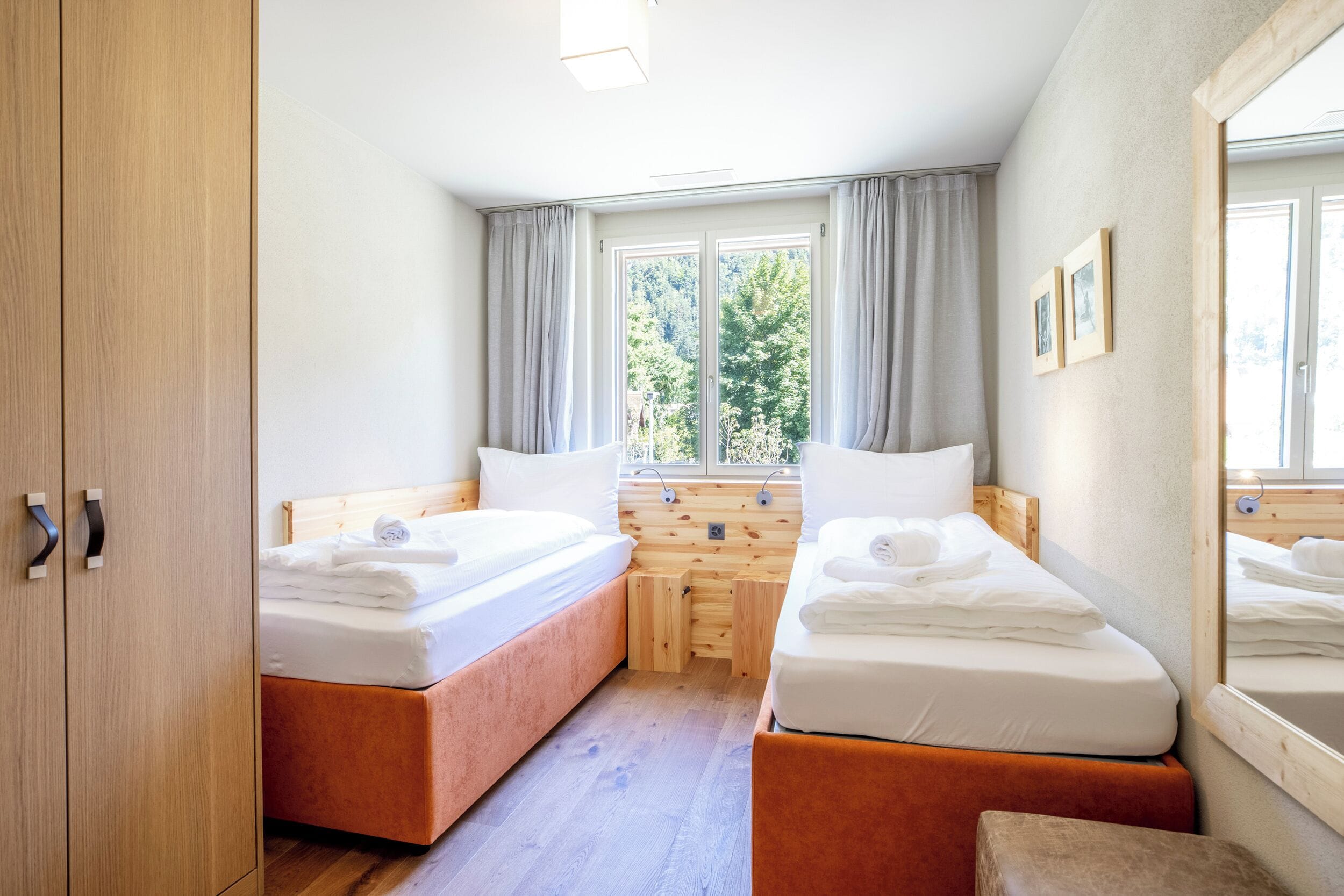 Una habitación con dos camas, un armario de madera, una ventana con cortinas y vistas a árboles.