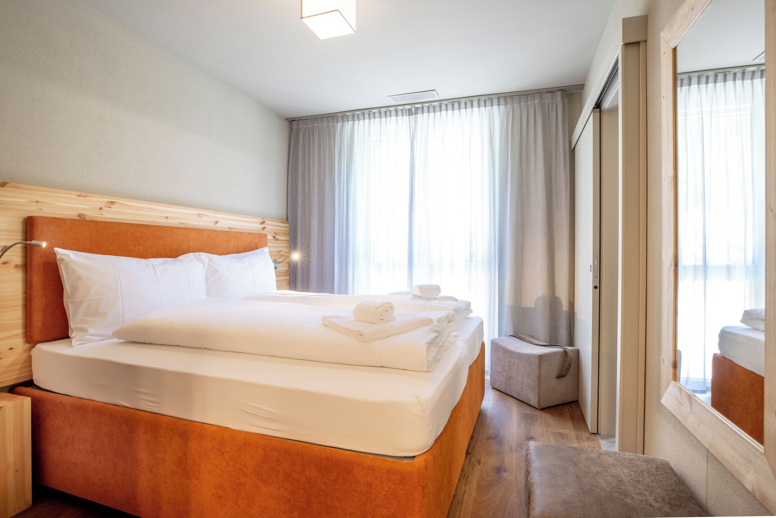 Habitación de hotel con cabecera de madera, una cama grande con ropa blanca, una mesita de noche y vistas al exterior a través de un ventanal.