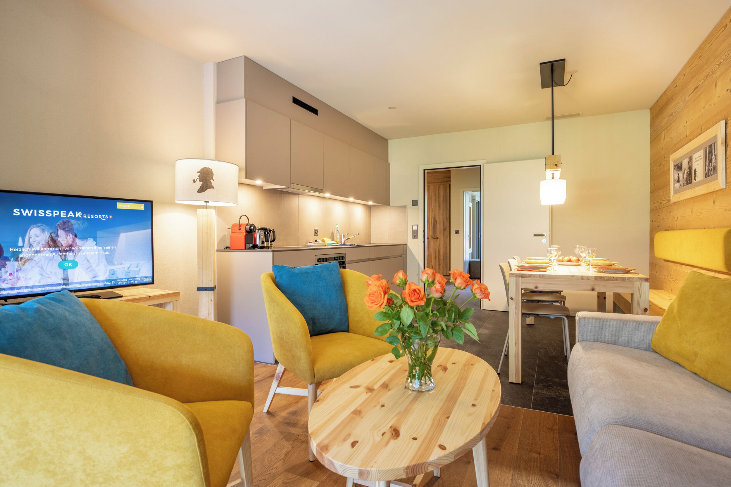A modern living room with a TV, yellow and grey sofas, a wooden coffee table with flowers, a kitchen in the background, and a dining area.