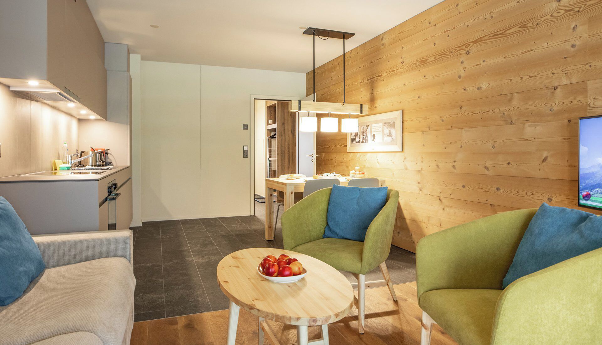 A modern living room with a dining area, wooden wall paneling, a television, and a bowl of fruit on a small table.